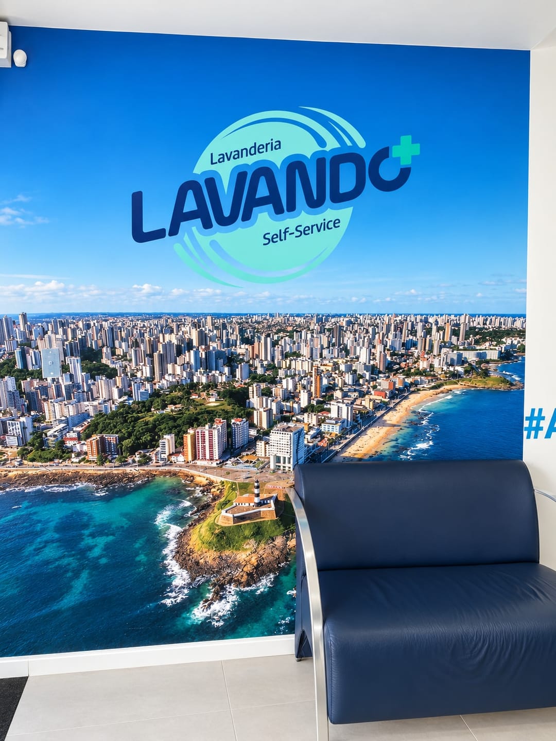 Mural com o logotipo da Lavando Mais sobre a vista aérea da Barra e do Farol, com uma poltrona azul-marinho à frente